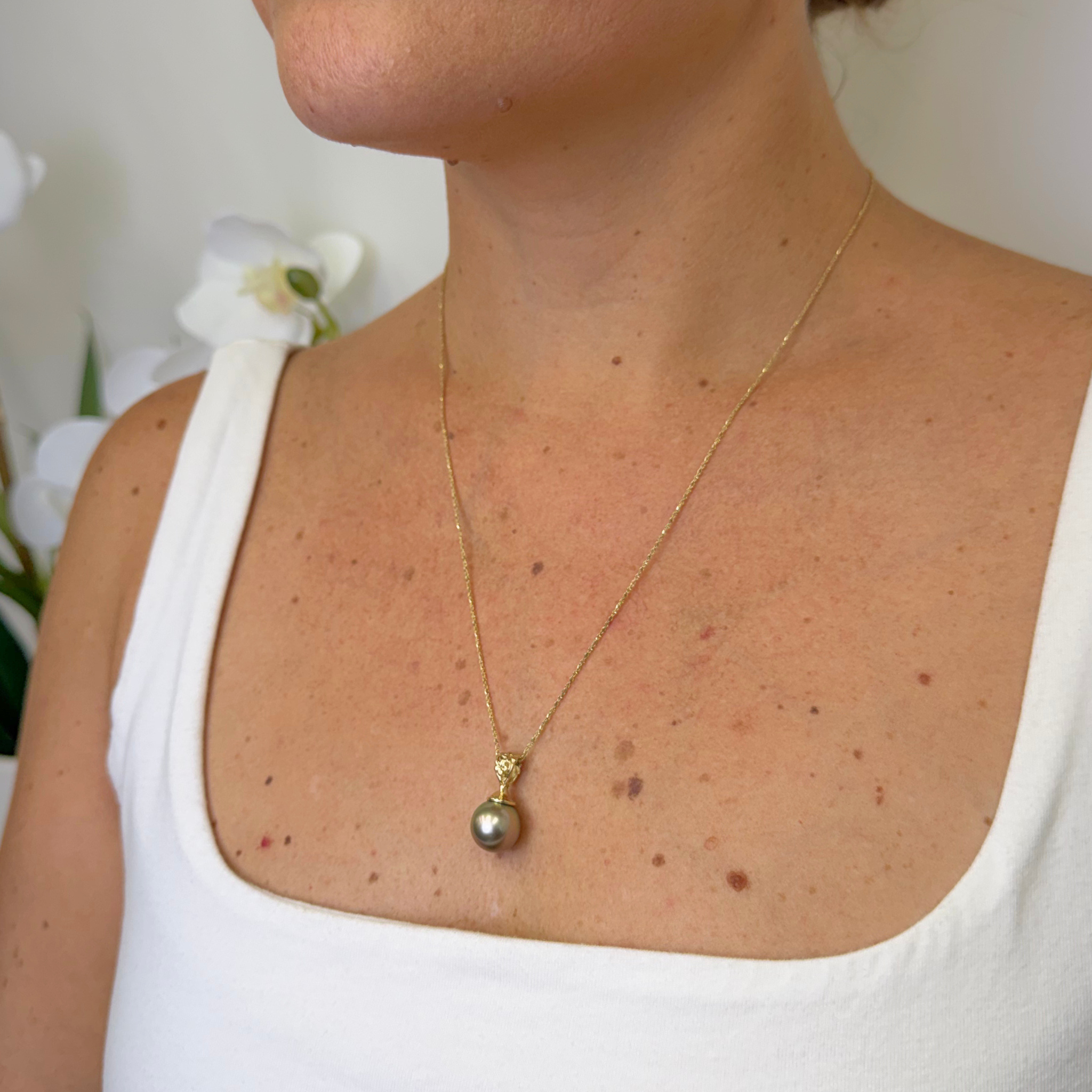 A close-up of a person wearing a delicate gold necklace with a single Tahitian pearl pendant, styled with a white tank top.