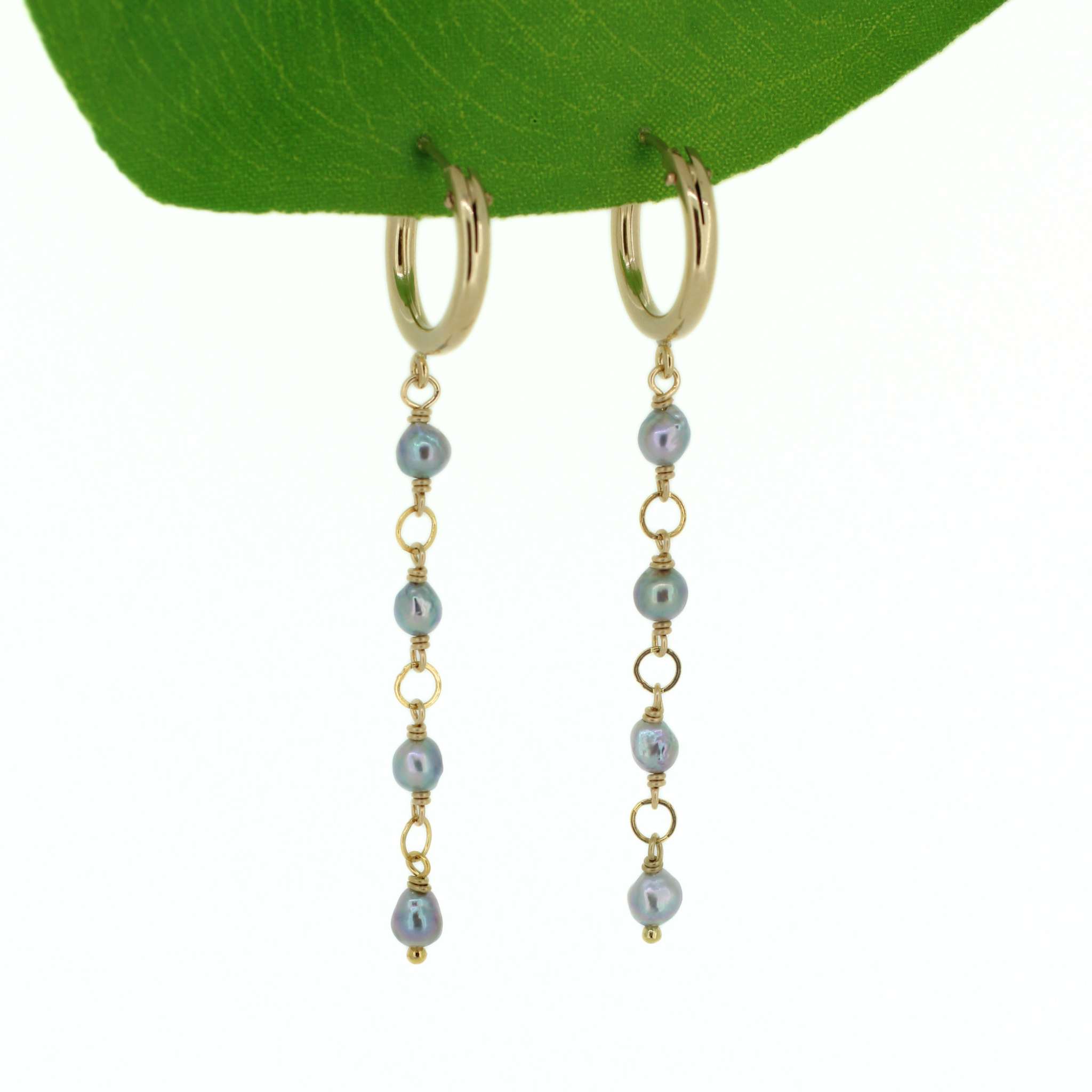 A pair of gold hoop dangle earrings with small iridescent blue-gray akoya pearls and gold spacer rings, hanging from a green leaf against a white background. Handmade by Brianne & Co. in Kauai, Hawaii