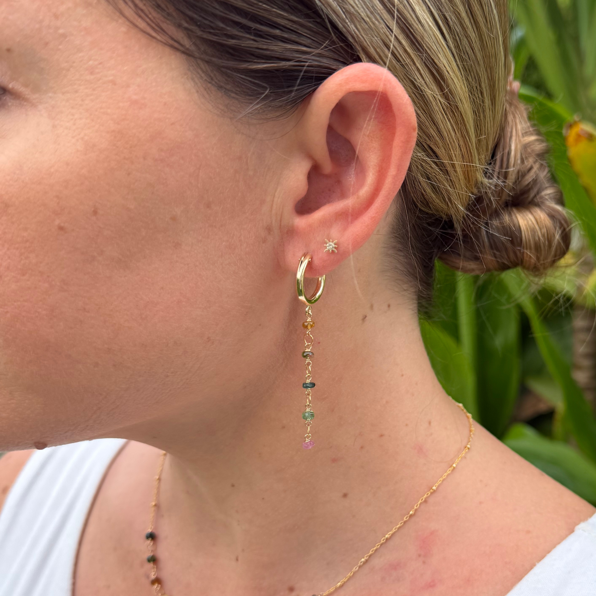 A close-up of a woman's ear wearing a gold huggie hoop earring with a dangling multicolored tourmaline beads against a blurred green outdoor background.