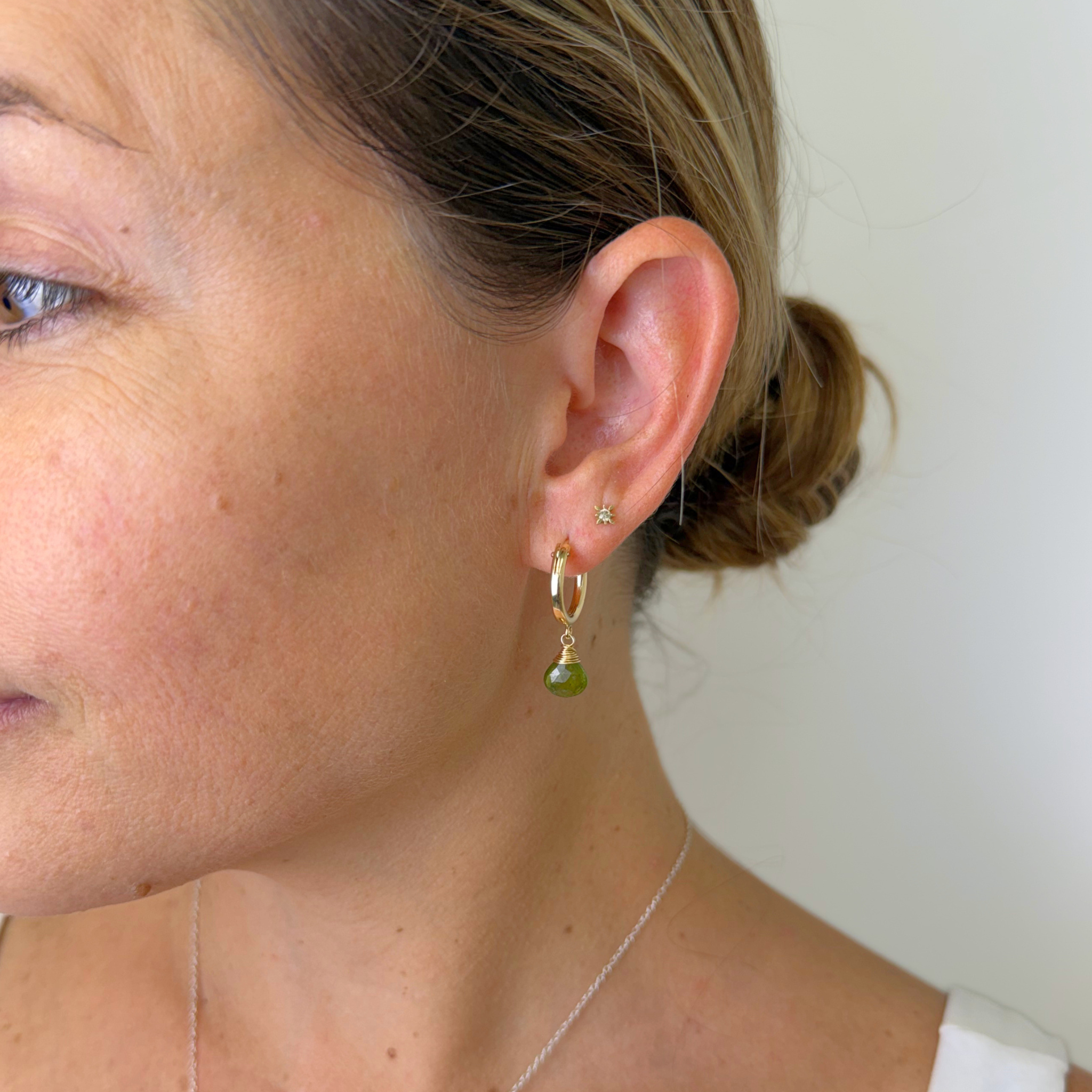 A close-up side profile of a woman wearing a delicate gold hoop earring with a small green gemstone drop and a tiny gold stud earring, against a plain light background.