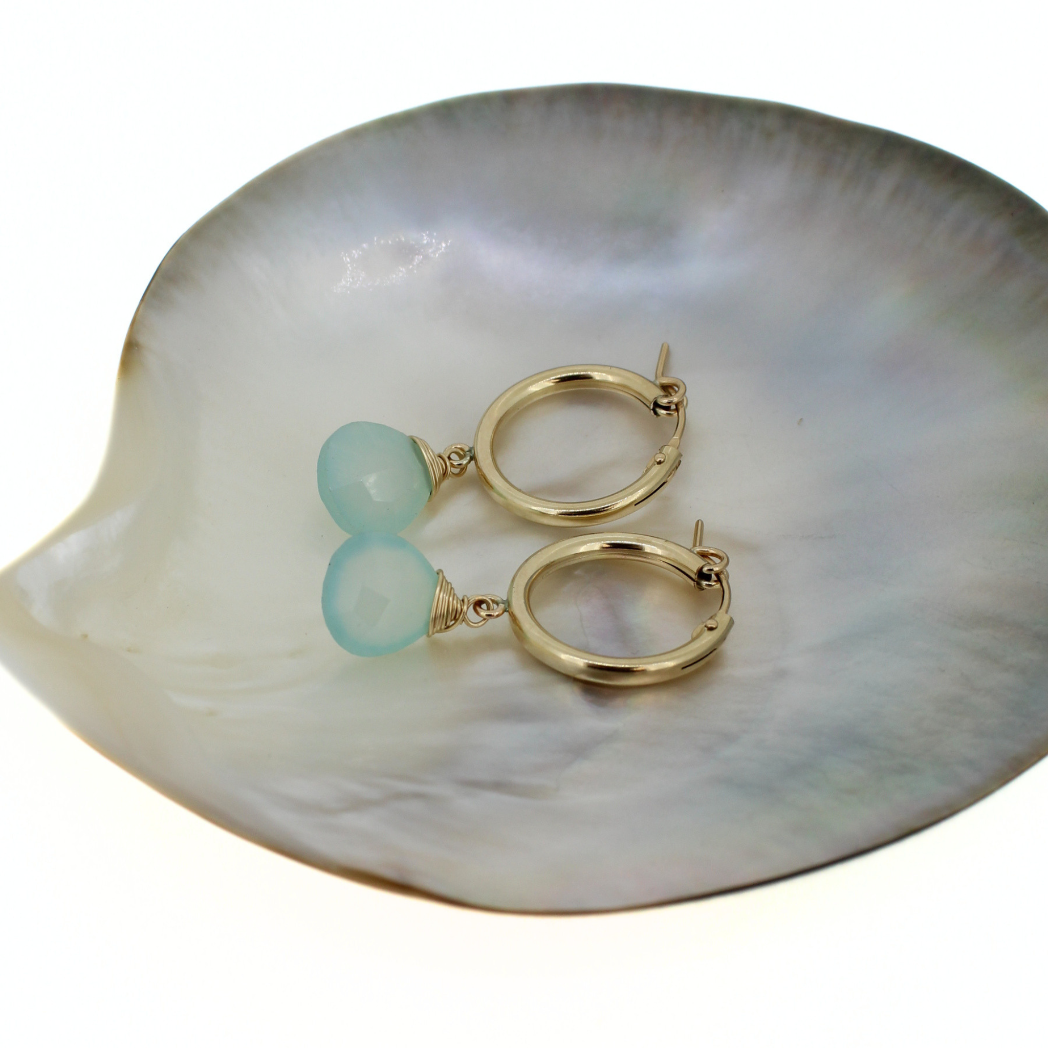 A pair of gold hoop earrings with light blue faceted blue chalcedony drops, displayed on a mother-of-pearl shell against a white background. Handmade by Brianne & Co. in Kauai, Hawaii