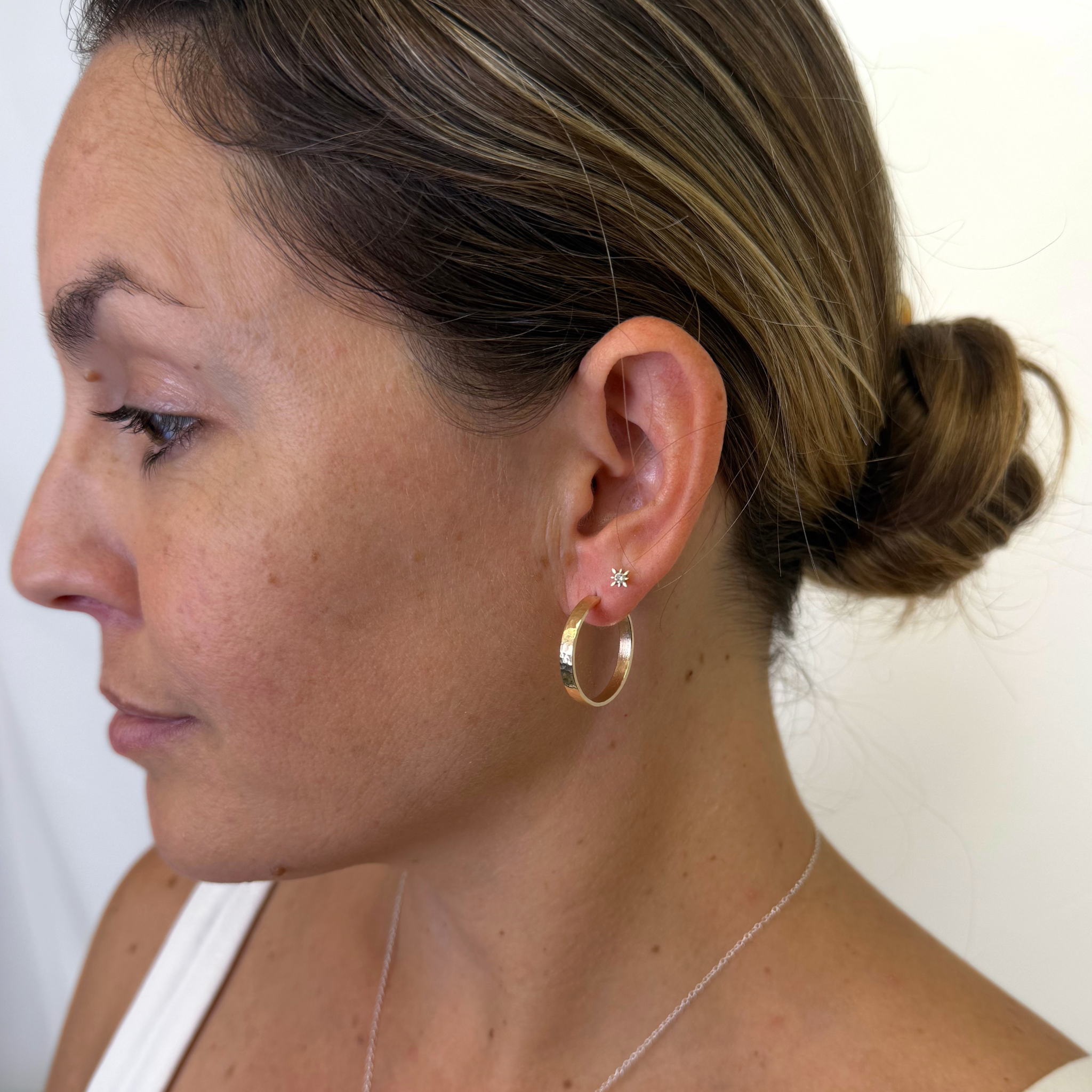 A close-up side profile of a woman wearing a polished gold hoop earring and a small star stud earring, with her hair in a low bun.