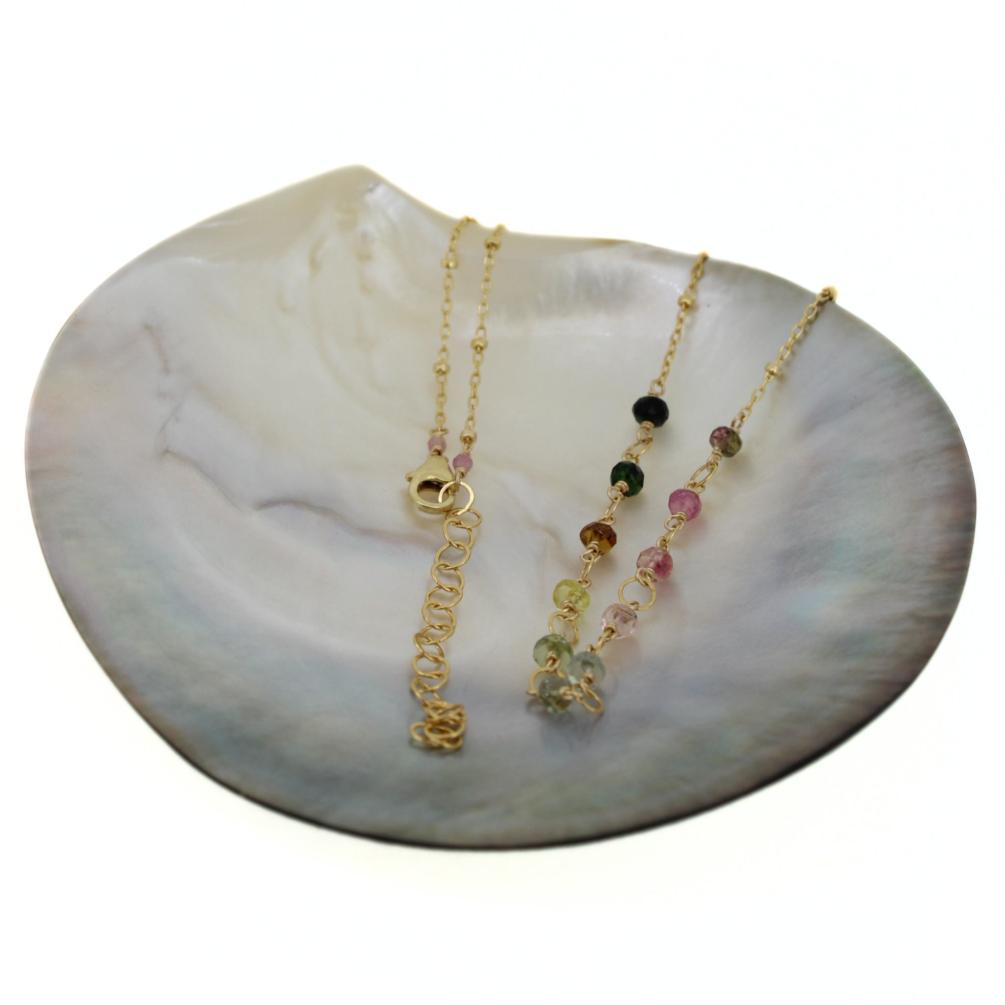 A delicate gold necklace displayed on a white mother-of-pearl shell dish against a white background.
