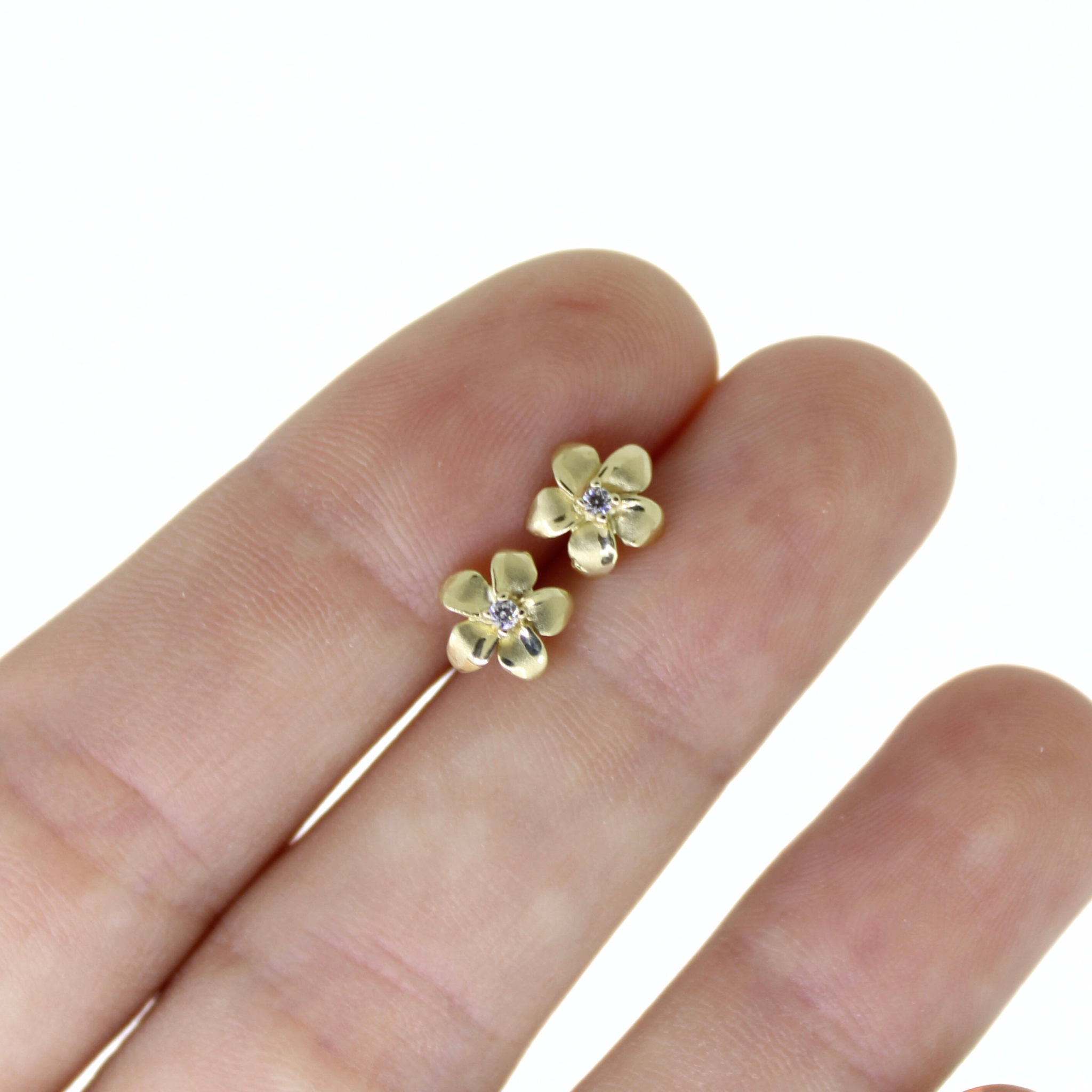 Brianne & Co. 14k yellow gold small diamond plumeria stud earrings between fingers