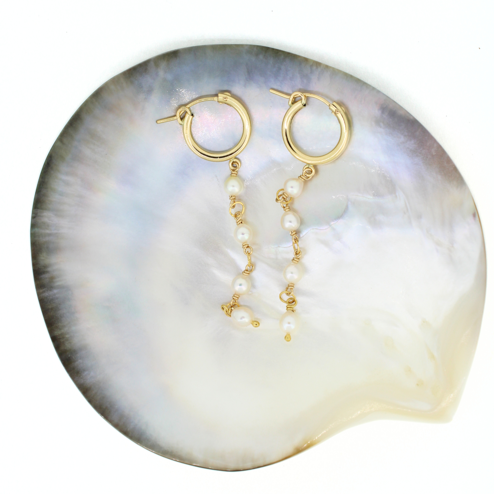 Gold hoop earrings with dangling pearl chains, displayed on a large iridescent shell against a white background.