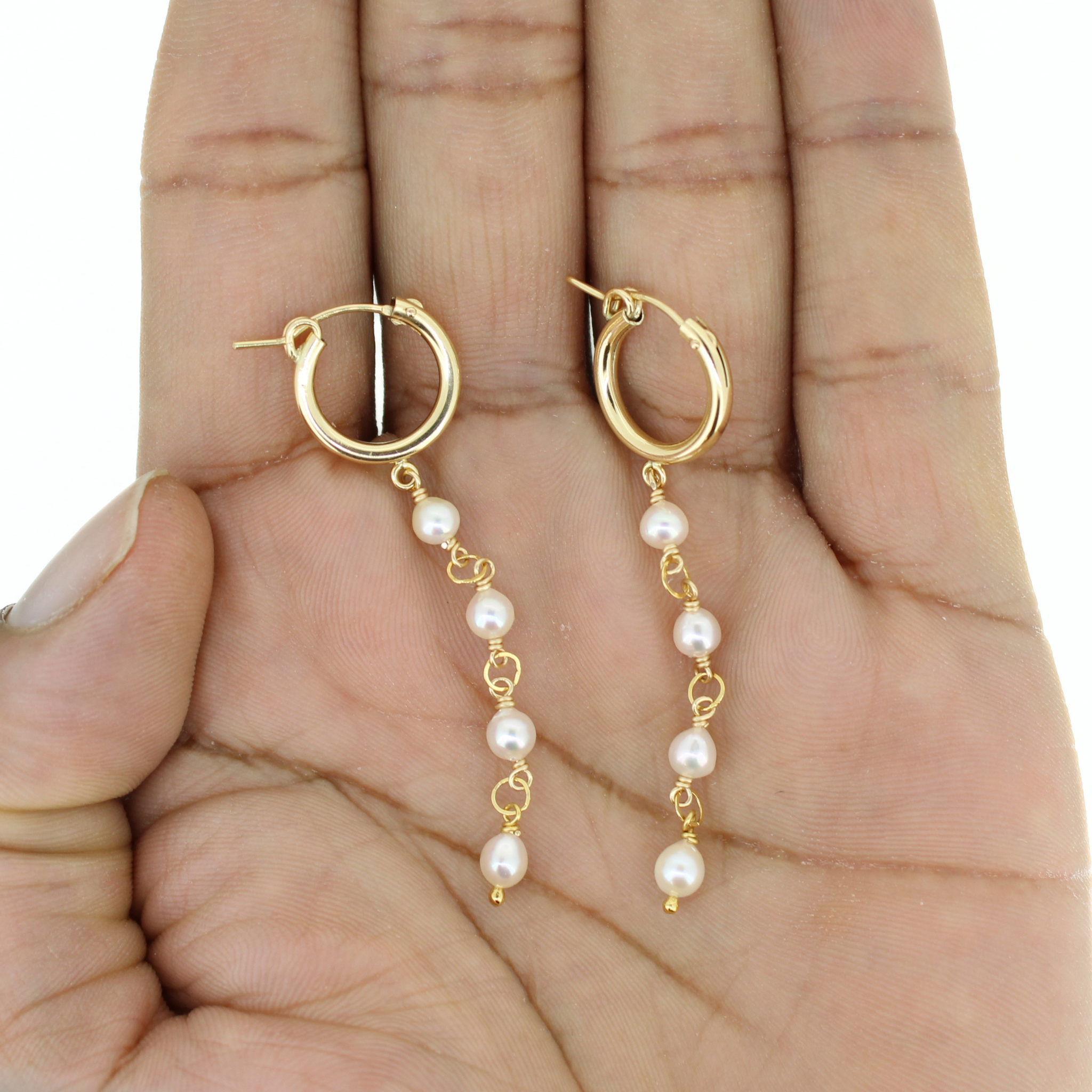 A pair of gold hoop dangle earrings with small white pearls, held in the palm of a hand.