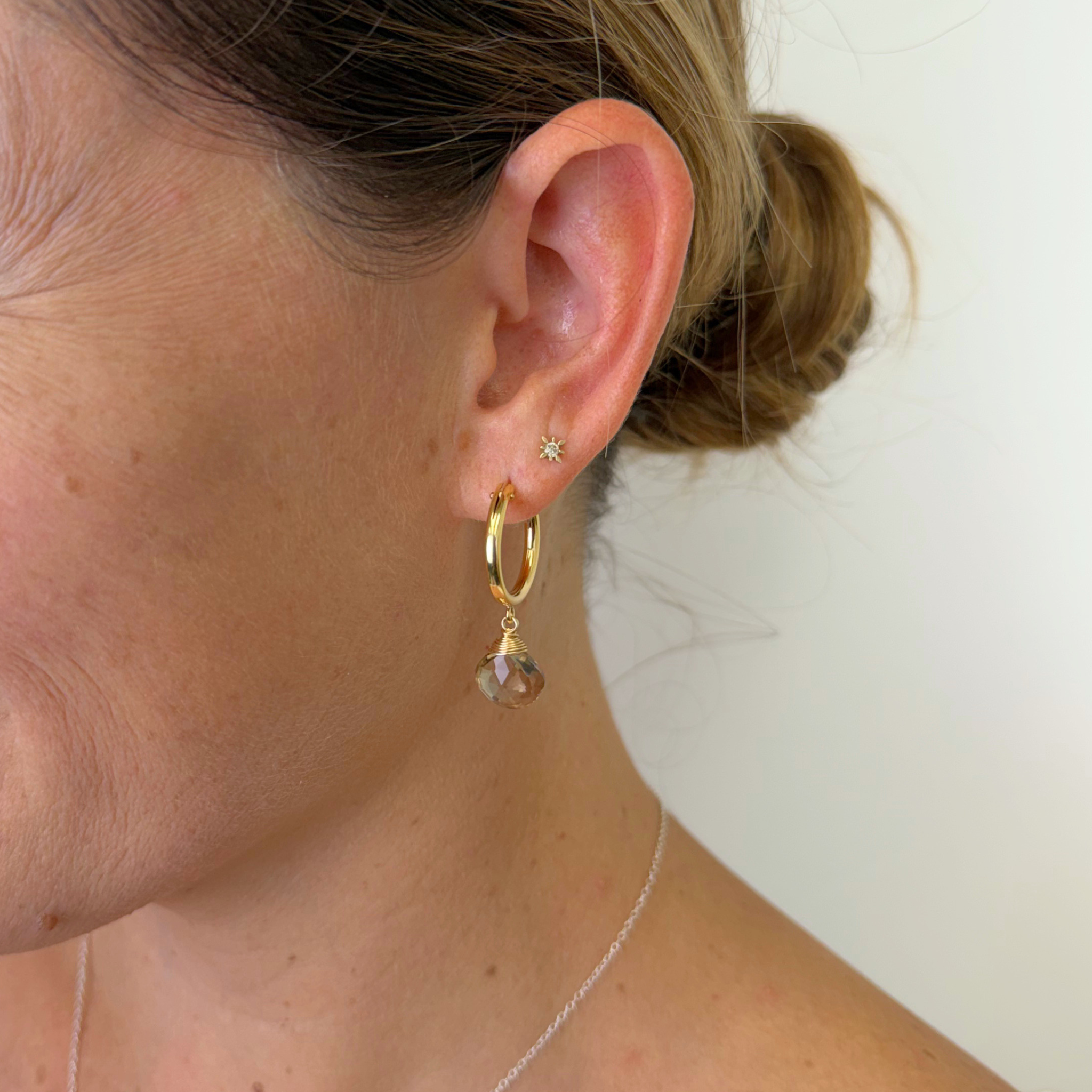 A close-up of a woman's ear wearing a delicate gold huggie hoop earring with a dangling faceted topaz gemstone, along with a small star stud earring.