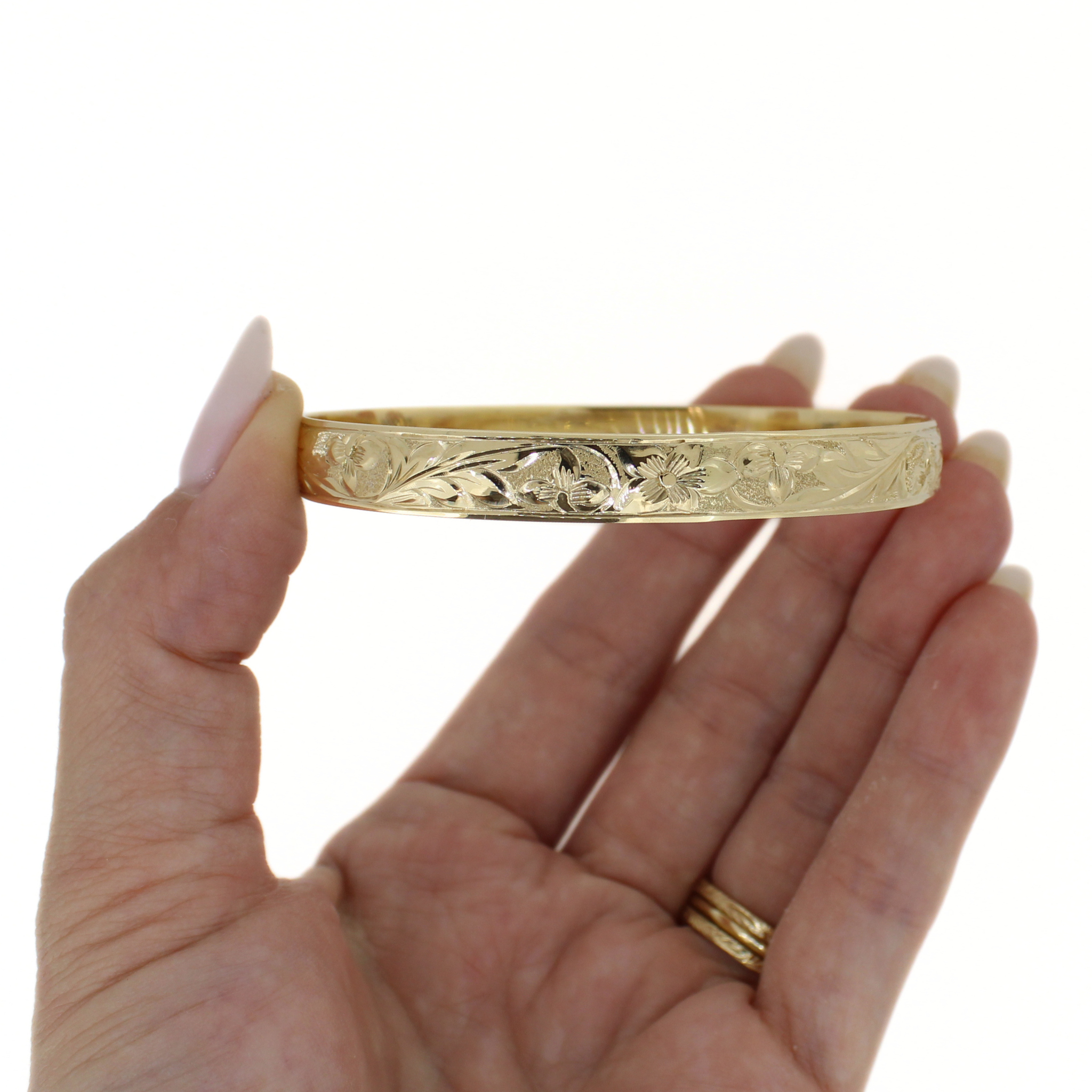 Gold bangle held sideways, with engraved tropical floral and wave detailing