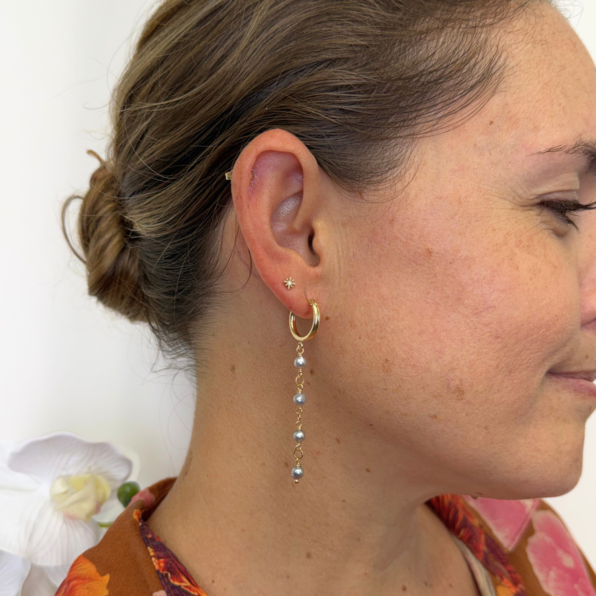 A close-up side profile of a woman wearing a delicate gold hoop earring with a dangling chain of small gray pearls, along with other small ear piercings.