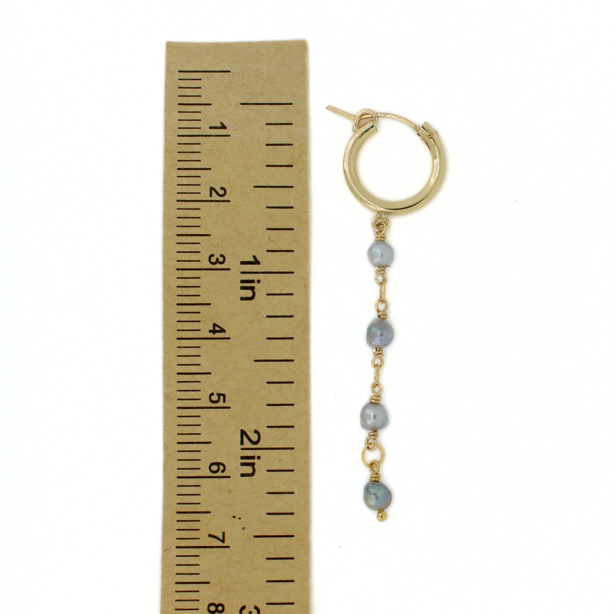 A gold hoop earring with a dangling chain of light blue-gray pearls and small gold spacers, shown next to a brown paper ruler for scale on a white background.