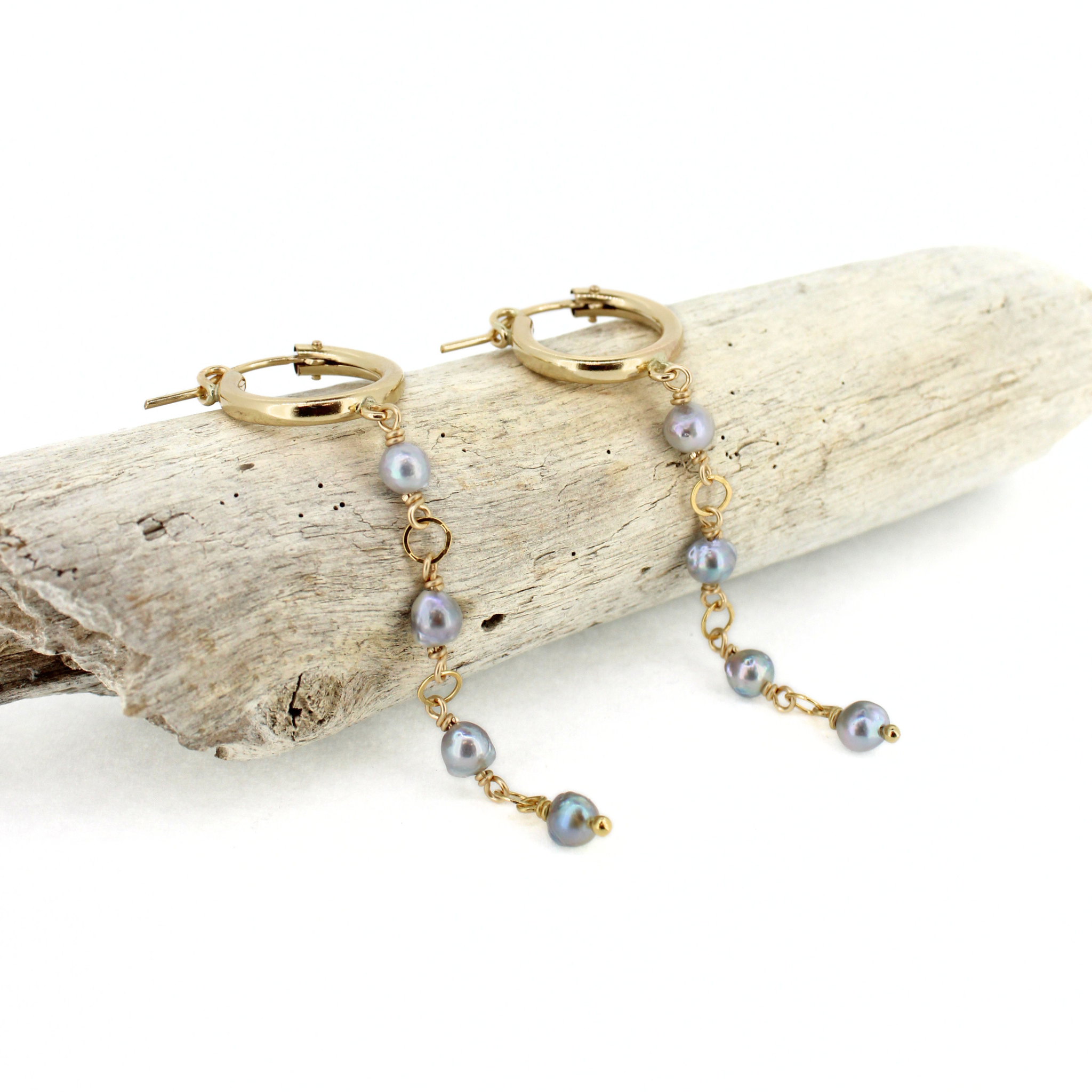 A pair of gold hoop dangle earrings with small gray pearls and gold chain links, displayed on a piece of driftwood against a white background.