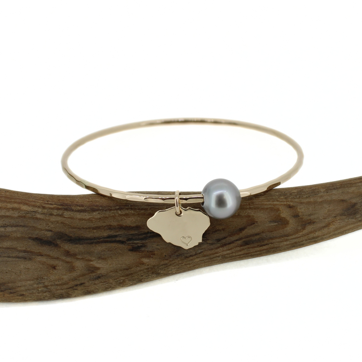 A gold bangle bracelet with a small Kauai-shaped charm and a gray pearl, displayed on a curved piece of wood against a white background.