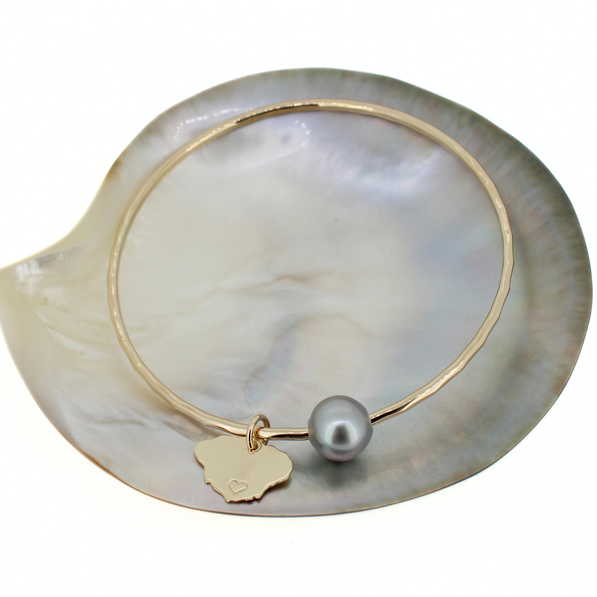 A gold bangle bracelet with a small gold Kauai island charm and a gray pearl, displayed on a white mother-of-pearl clam shell against a white background.