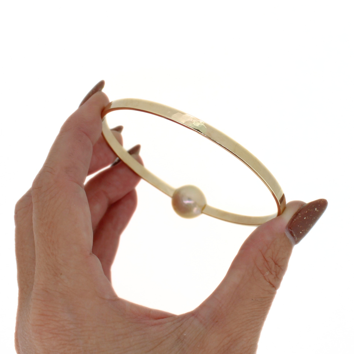 A gold bangle bracelet with a small white pearl, held by a hand with glittery brown nail polish against a solid white background.