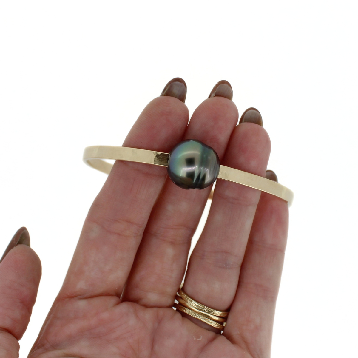 A gold bangle bracelet with a central iridescent green-black Tahitian pearl, held on a person's fingers against a plain white background.