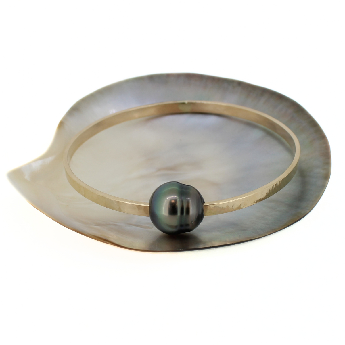 A thick hammered gold bangle bracelet featuring a single dark gray-black Tahitian pearl, displayed on a mother-of-pearl shell dish against a white background.