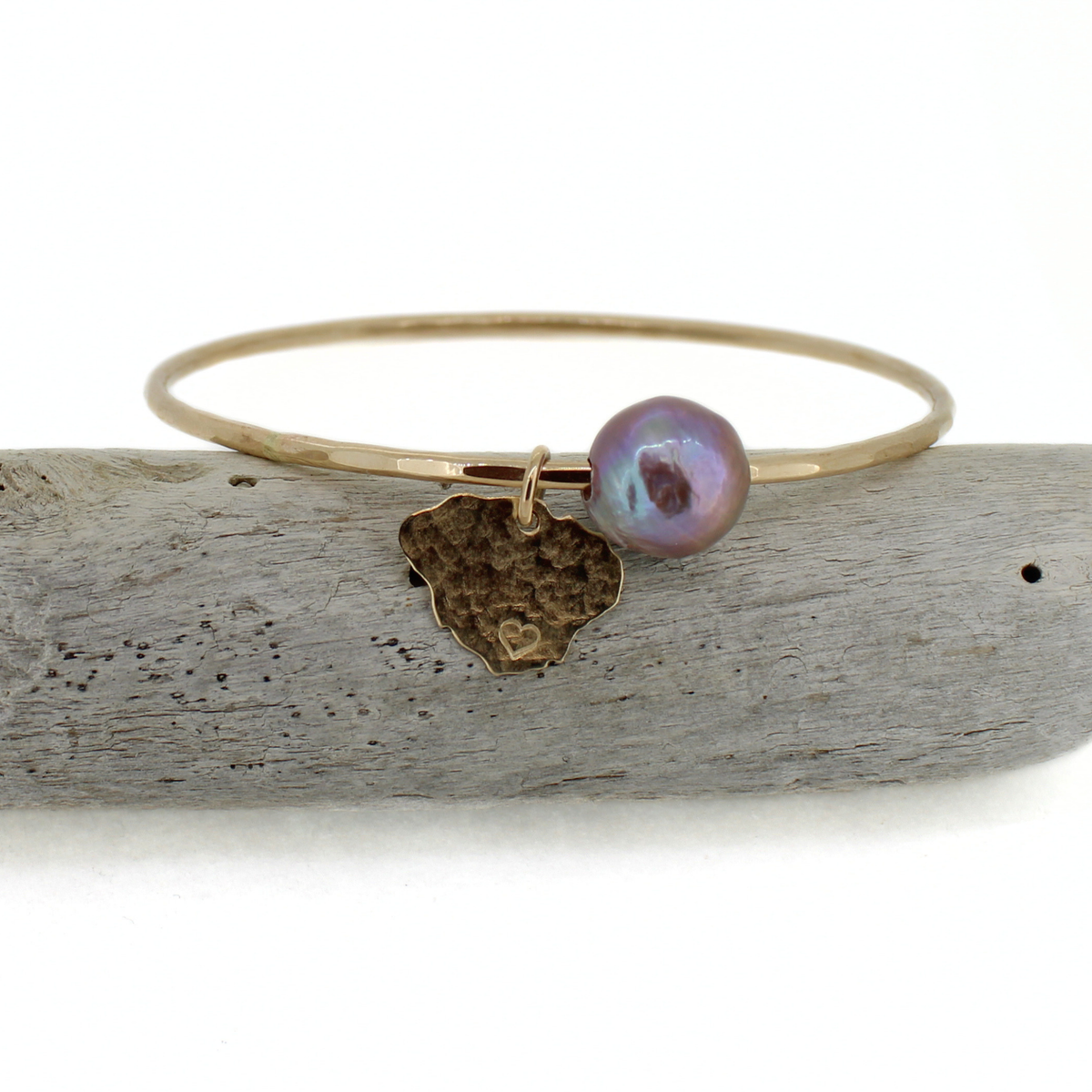 A gold bangle bracelet with a hammered Kauai charm and a baroque iridescent pearl, displayed on a piece of weathered driftwood against a white background.