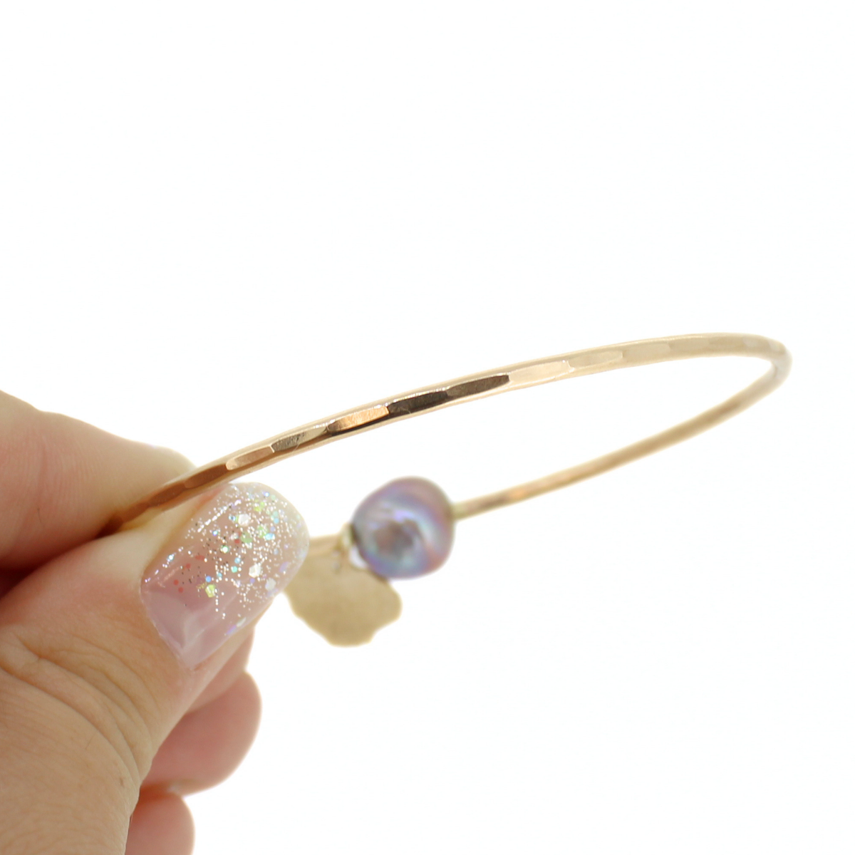 A gold bangle bracelet with a small iridescent pearl and a tiny gold charm, held by a hand with glittery nail polish against a white background.
