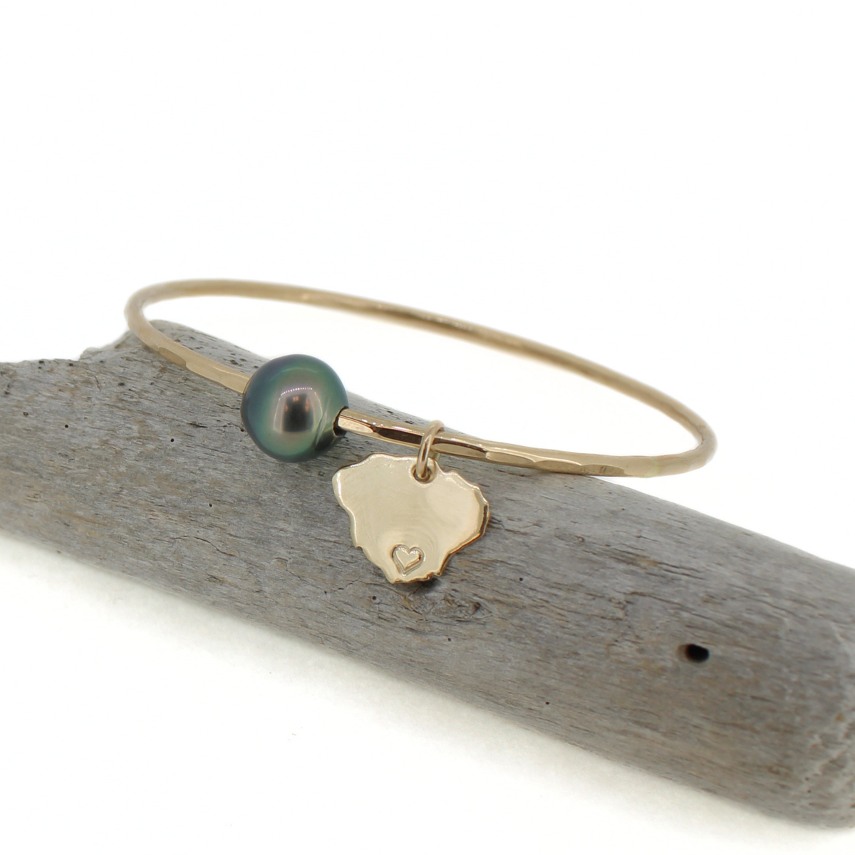 A delicate gold bangle bracelet with a dark green iridescent Tahitian pearl and a small gold Kauai island charm, displayed on a piece of weathered driftwood against a white background.