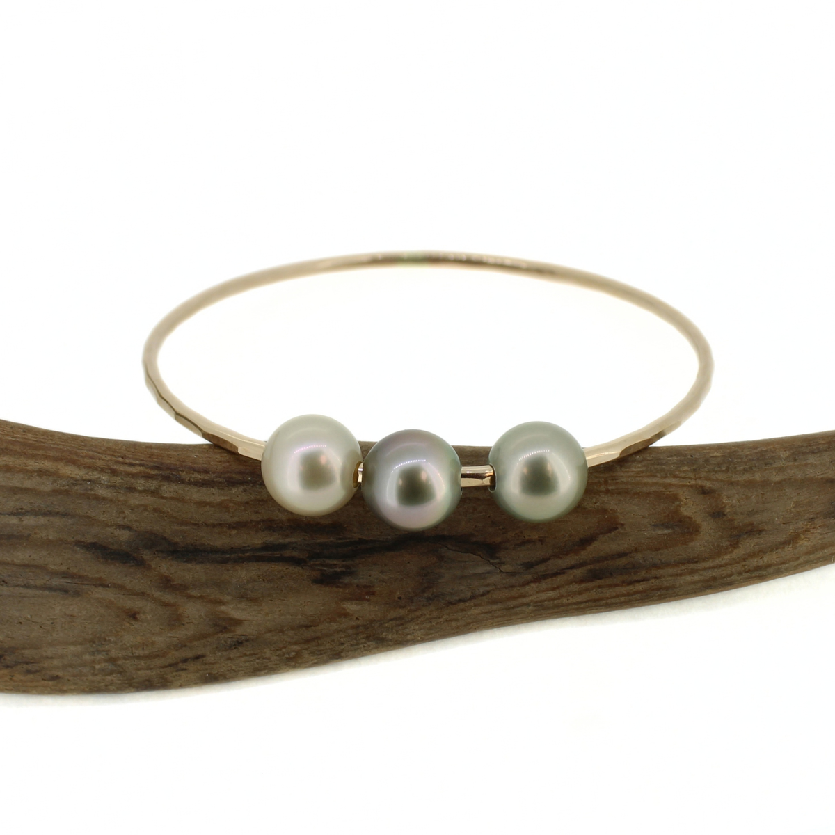 A thin gold bangle bracelet featuring three round Tahitian pearls in shades of gray, lavender, and green, displayed on a curved piece of dark wood against a white background.