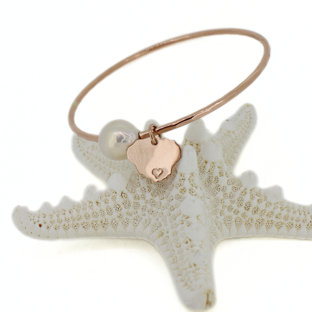 A rose gold bangle bracelet with a small Kauai charm, and a white pearl bead, displayed on a white starfish prop against a white background.