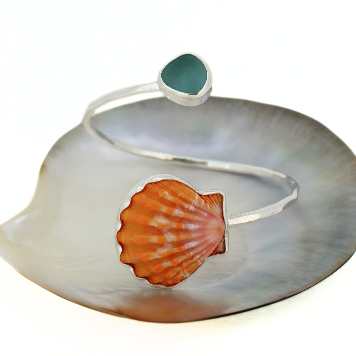 A silver open cuff bracelet featuring a large orange sunrise shell on one side and a small round blue sea glass on the other, displayed on a white shell-shaped prop.