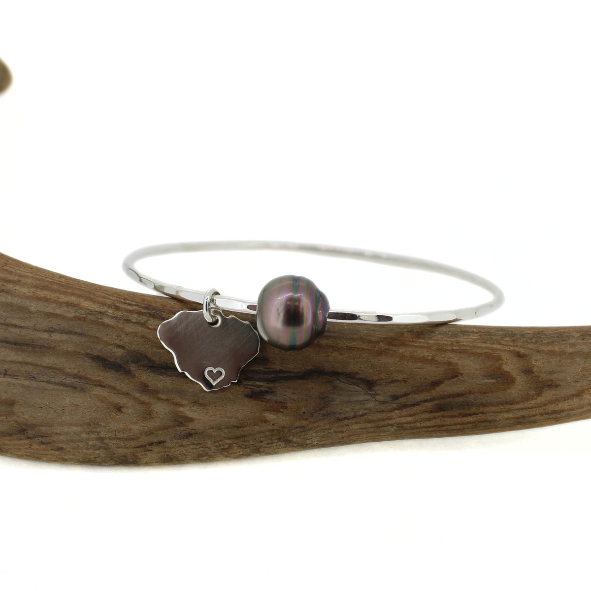 Silver hammered bangle bracelet with a dark Tahitian pearl and Kauai island charm displayed on a piece of driftwood
