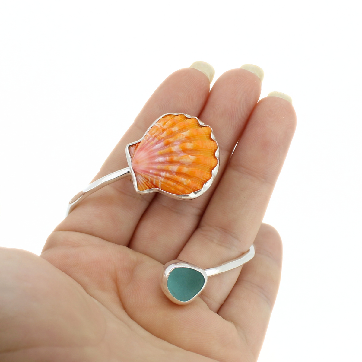 A hand holding a silver sunrise shell and sea glass cuff bracelet against a white background.