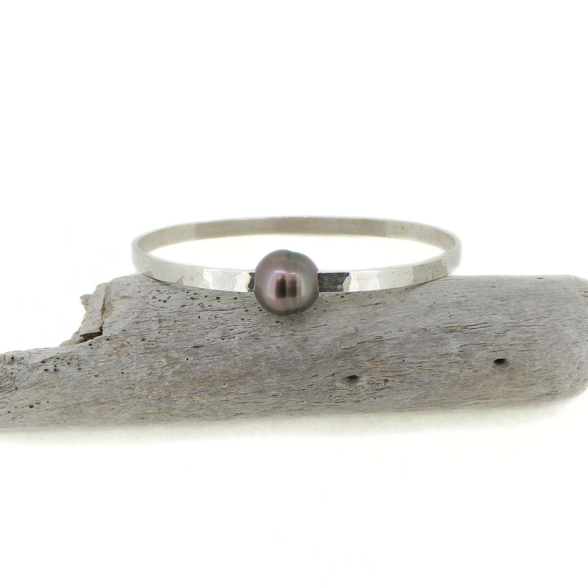 A minimalist sterling silver bangle bracelet with a single dark gray pearl, displayed on a piece of weathered driftwood against a white background.