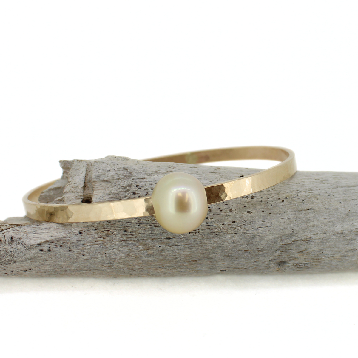 A hammered gold bangle bracelet with a single round golden south sea pearl, displayed on a piece of weathered driftwood against a white background.