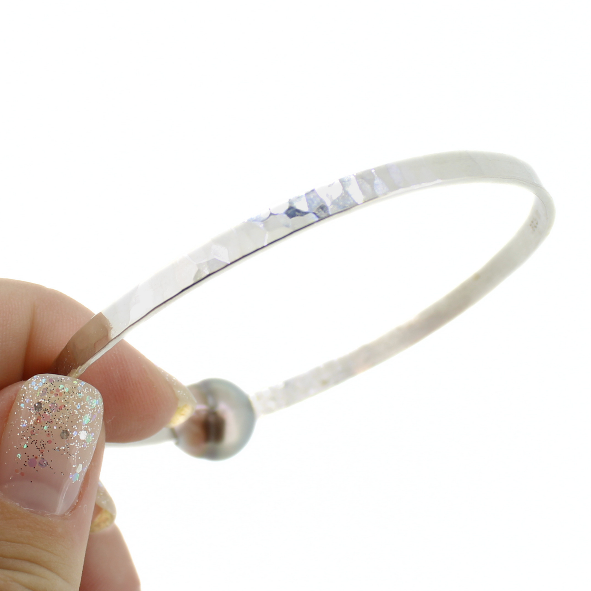 A silver hammered metal bangle bracelet held by a hand with glittery nail polish against a plain white background.