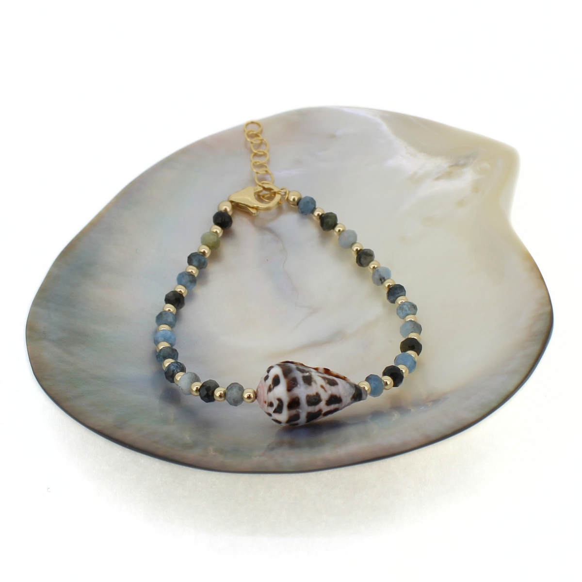 A beaded bracelet with a central spotted seashell at the center, displayed on a large mother-of-pearl shell dish. The bracelet features small faceted beads in blue and green, accented with gold spacers and a gold clasp.