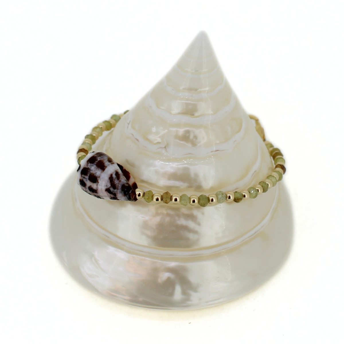 A delicate beaded bracelet with olive green and gold beads, featuring a small brown and white patterned shell accent, displayed on a white conical seashell stand against a white background.