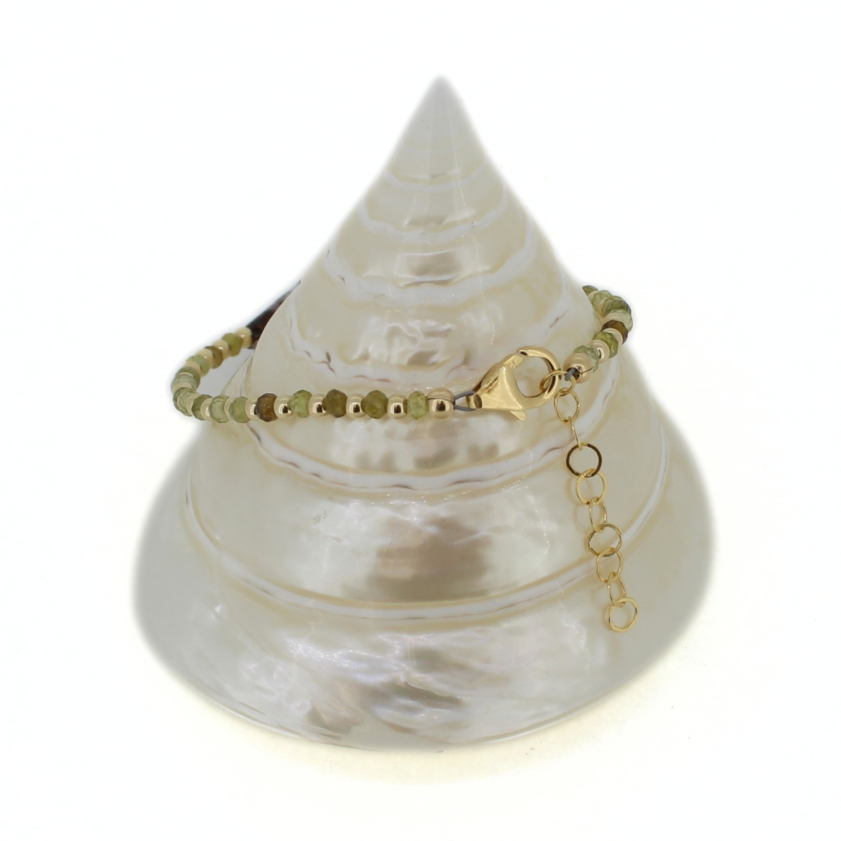 A delicate beaded bracelet with olive green beads and gold metal accents, displayed on a white seashell jewelry stand against a white background.