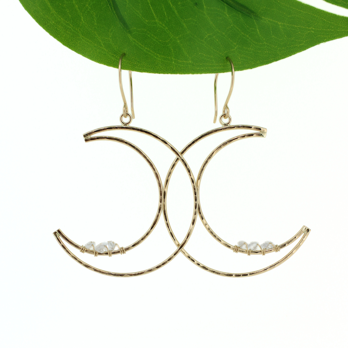 A pair of gold crescent moon dangle earrings with small clear Herkimer diamond accents, hanging from a green leaf against a white background.