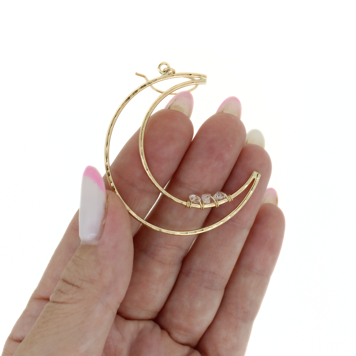 A pair of gold crescent hoop earrings with three small clear gemstone accents, held on a person's fingers against a plain white background.