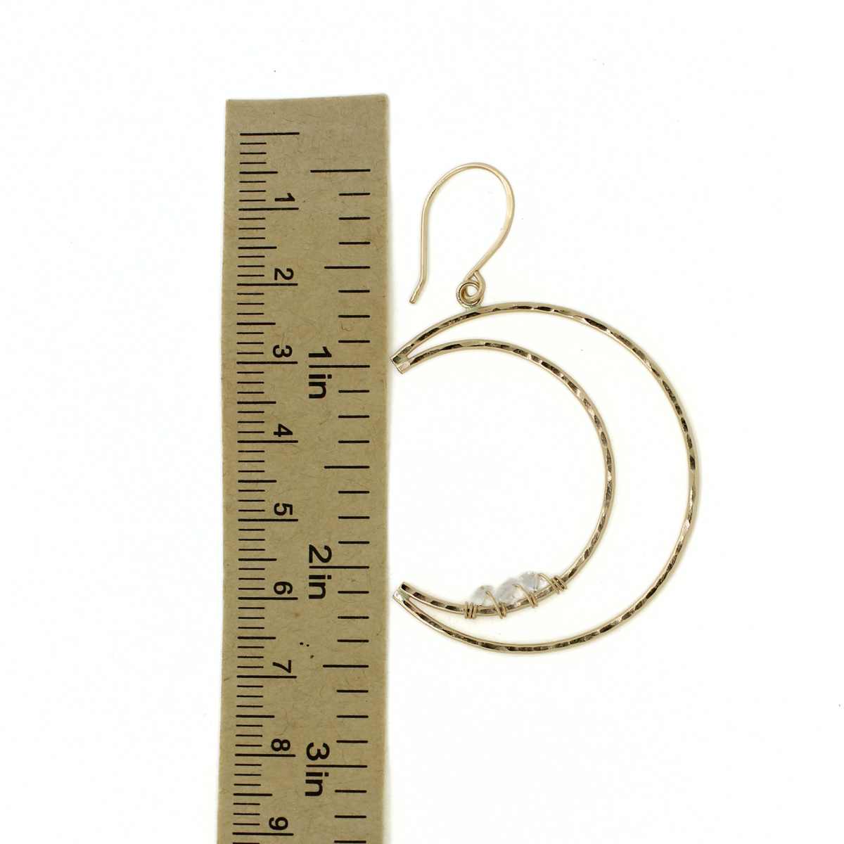 A pair of gold hoop earrings with a textured finish and a small clear crystal accent, shown next to a cardboard ruler for scale on a white background.