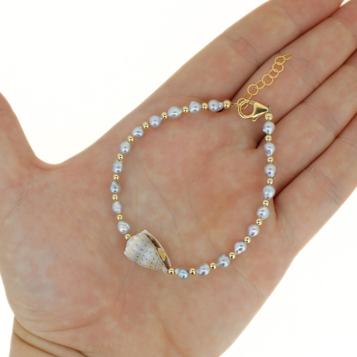 A delicate bracelet with small light blue pearls, gold spacer beads, and a natural seashell charm, displayed on an open hand against a white background.