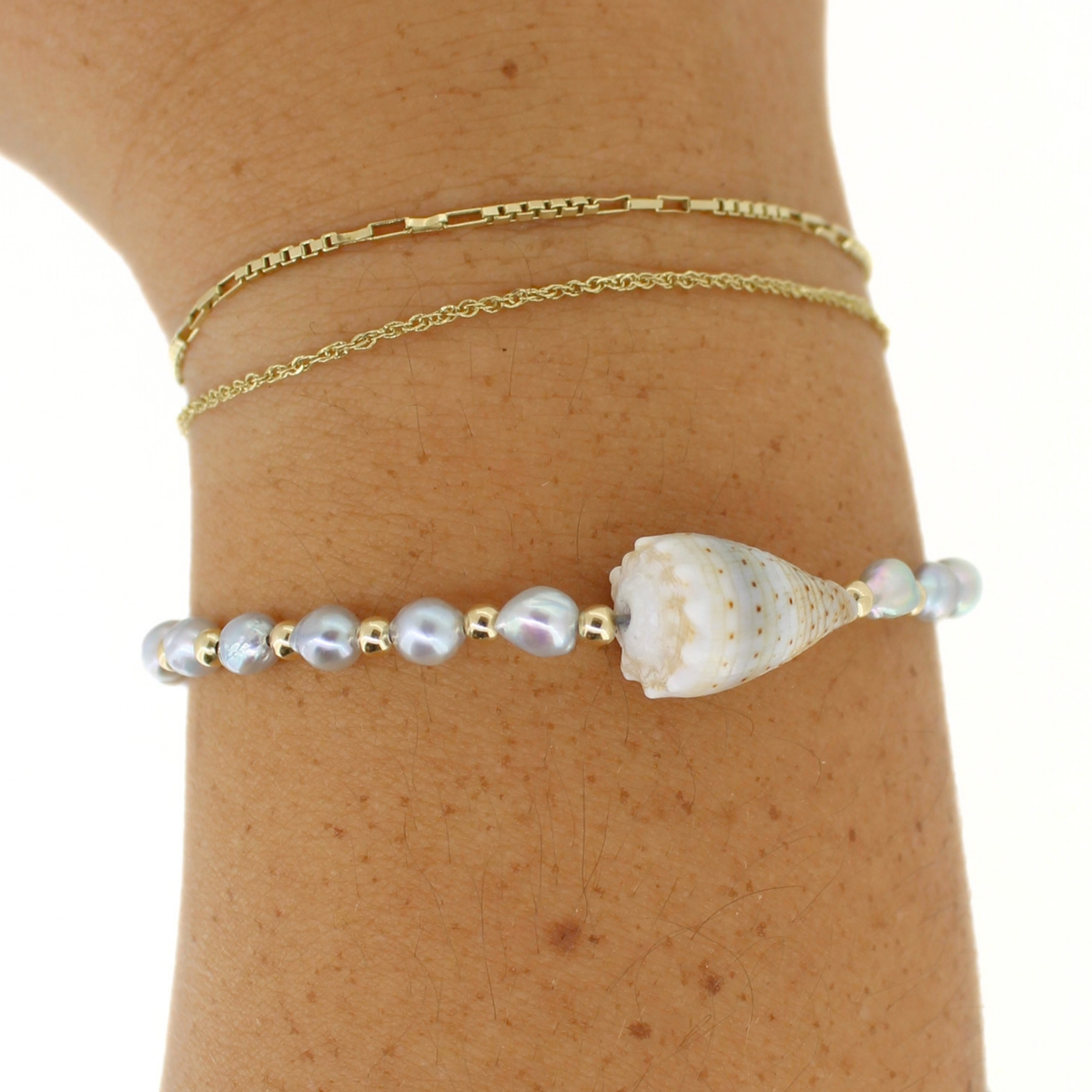 A close-up of a person's wrist wearing three delicate bracelets: a gold chain link bracelet, a thin gold chain bracelet, and a beaded bracelet featuring light blue baroque pearls, gold spacer beads, and a white seashell centerpiece.