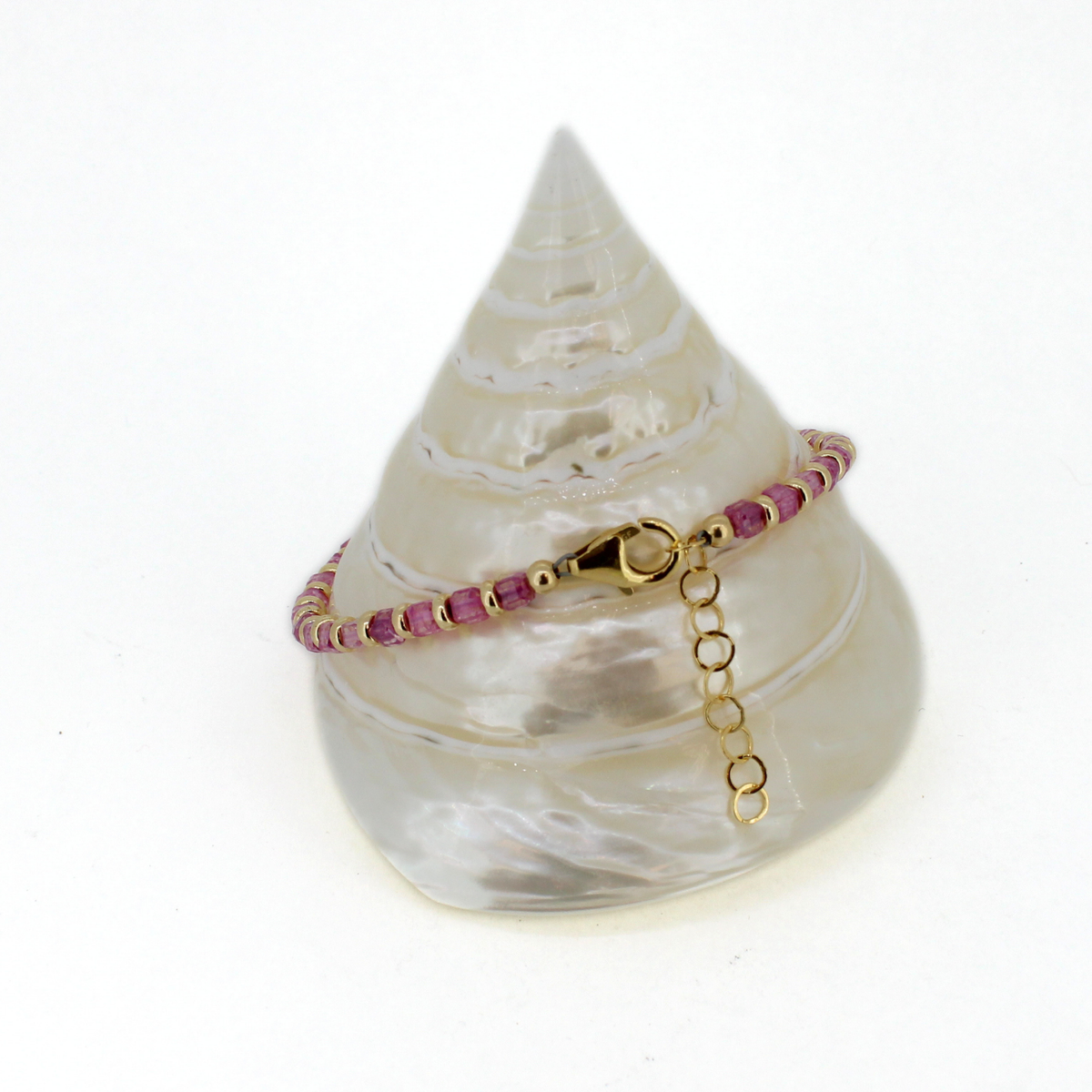 A delicate beaded bracelet with alternating pink and gold beads, featuring a gold clasp and extension chain, displayed on a white seashell stand against a white background.