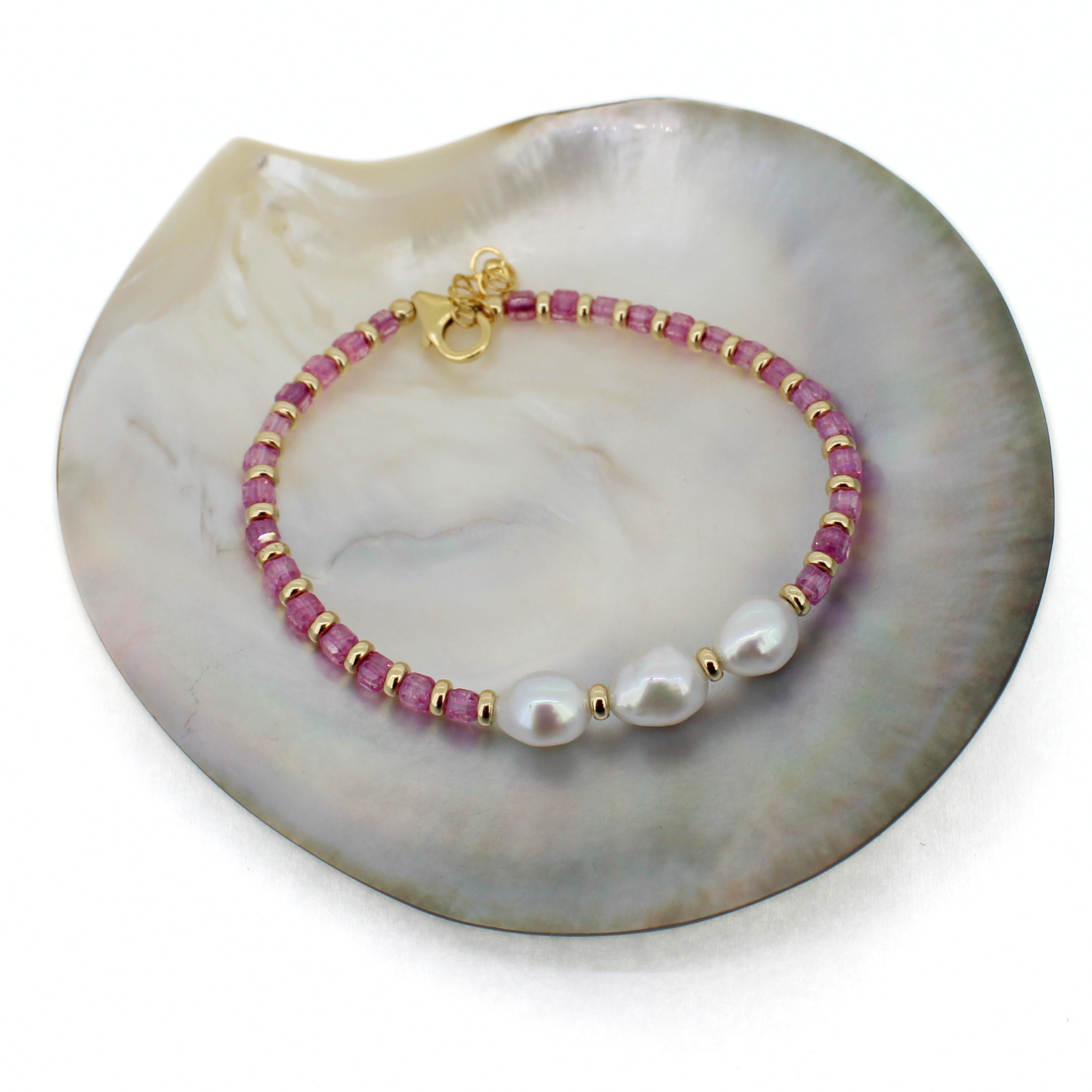 A beaded bracelet with pink cube-shaped beads, small gold spacer beads, and three white baroque pearls, displayed on a white mother-of-pearl shell.