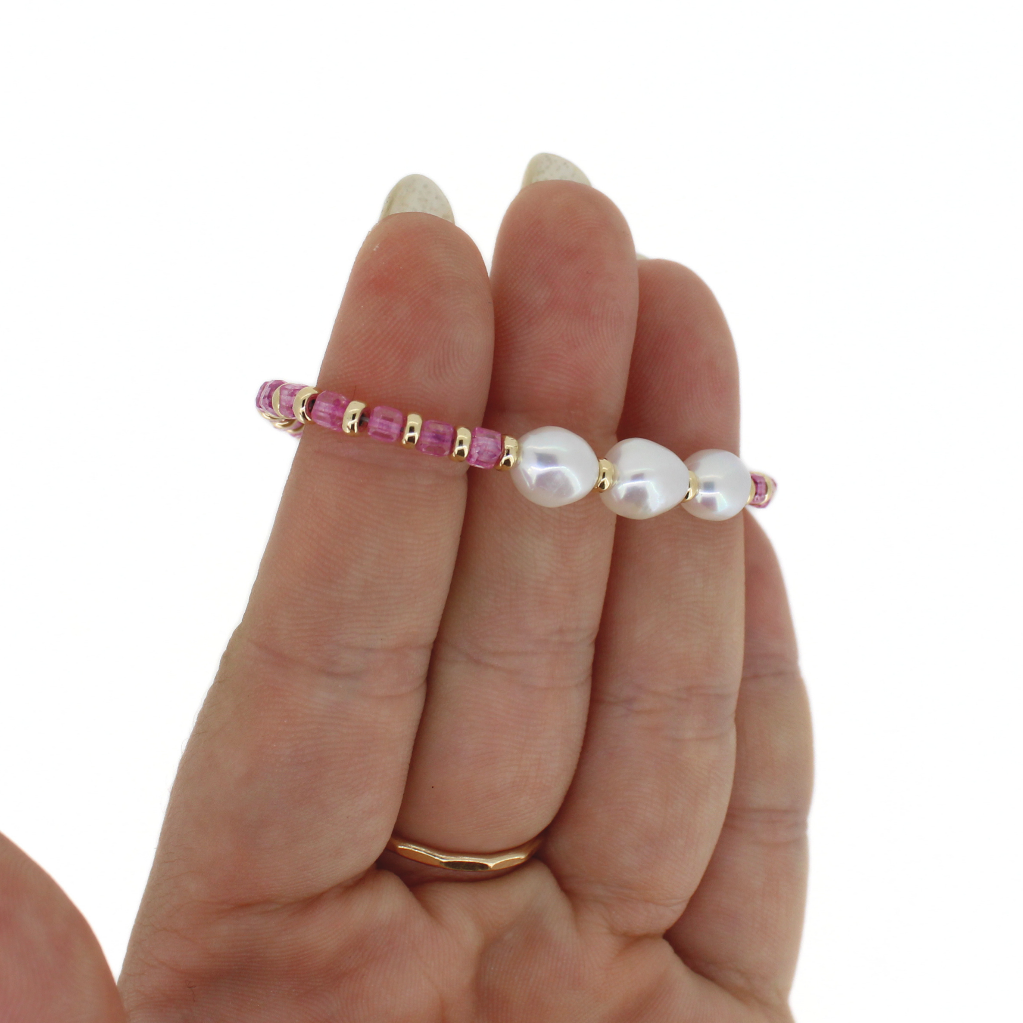 A delicate beaded bracelet with pink faceted beads, gold spacer beads, and three large white freshwater pearls, shown worn on a person's finger against a white background.