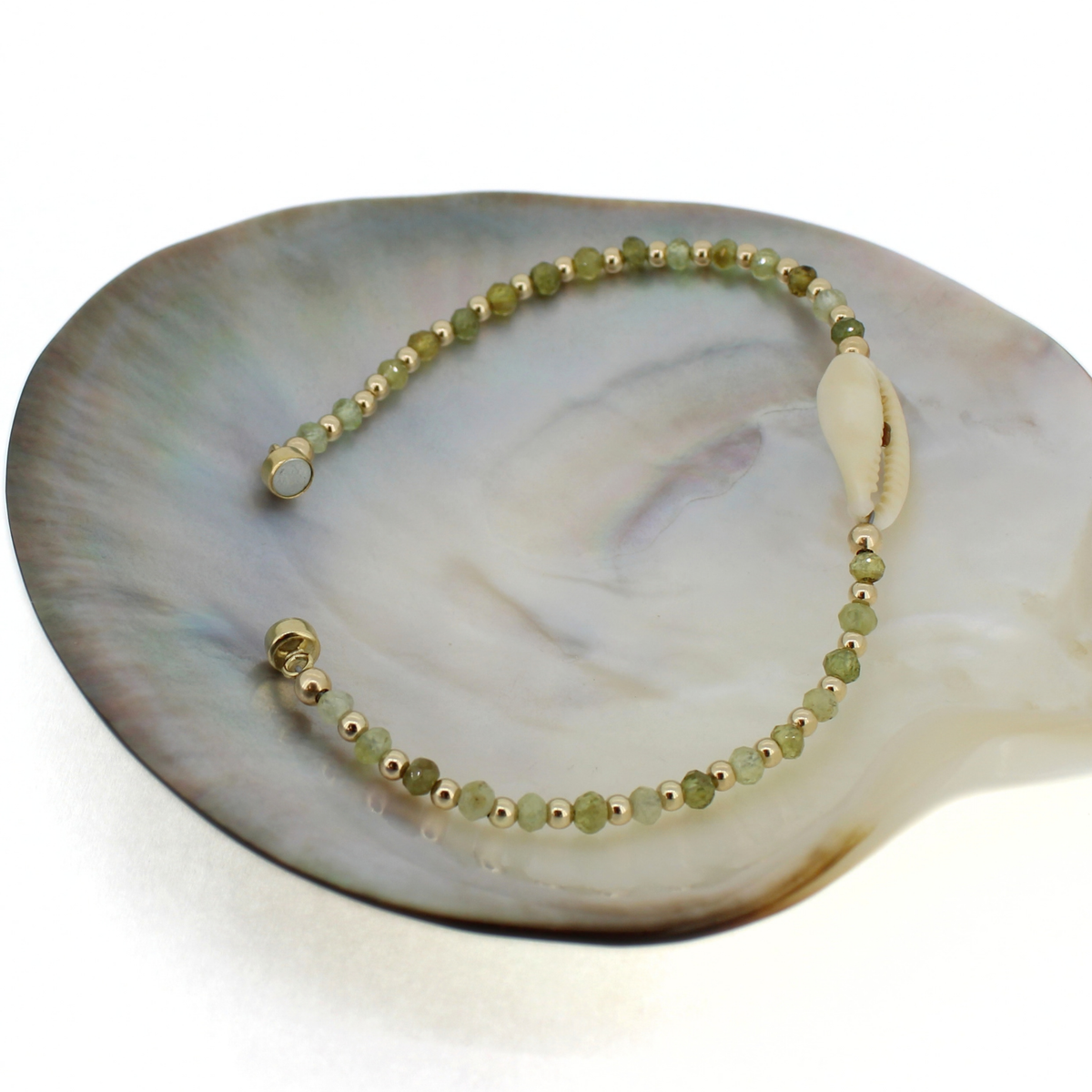 A beaded bracelet with light green faceted beads, small gold spacer beads, and a white cowrie shell, displayed on a large iridescent shell fragment.