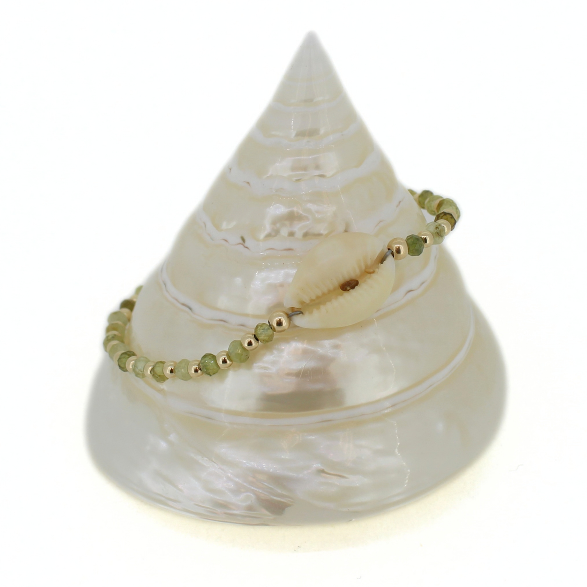 A delicate beaded bracelet with green beads and small gold spacer beads, featuring a central white cowrie shell, displayed on a white conical seashell stand against a white background.