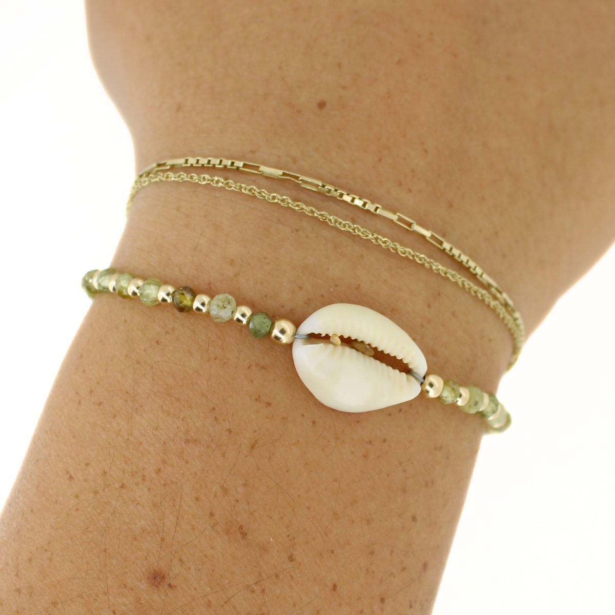A close-up of a person's wrist wearing a double-layered bracelet. The top layer is a delicate gold chain, and the bottom layer features a gold bracelet with small green beads and a central white cowrie shell.