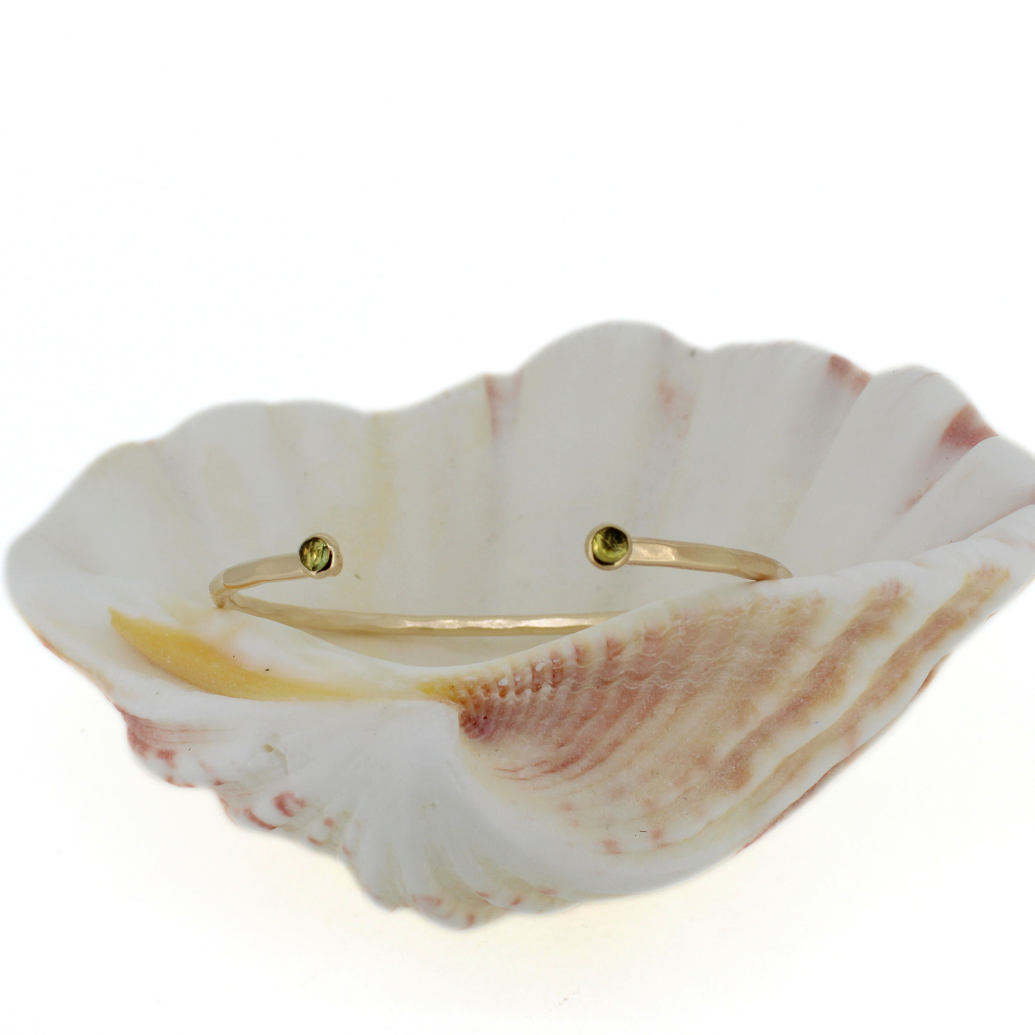 A delicate gold bangle bracelet with two small round green gemstone accents, displayed inside a white and pink seashell on a clean white background.