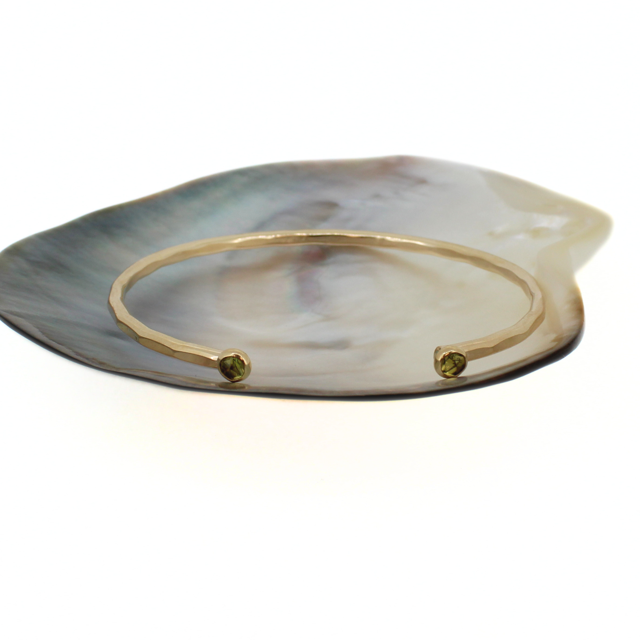 A gold-tone open cuff bangle bracelet with small green peridot gemstone accents at each end, displayed on a polished mother-of-pearl shell against a white background.