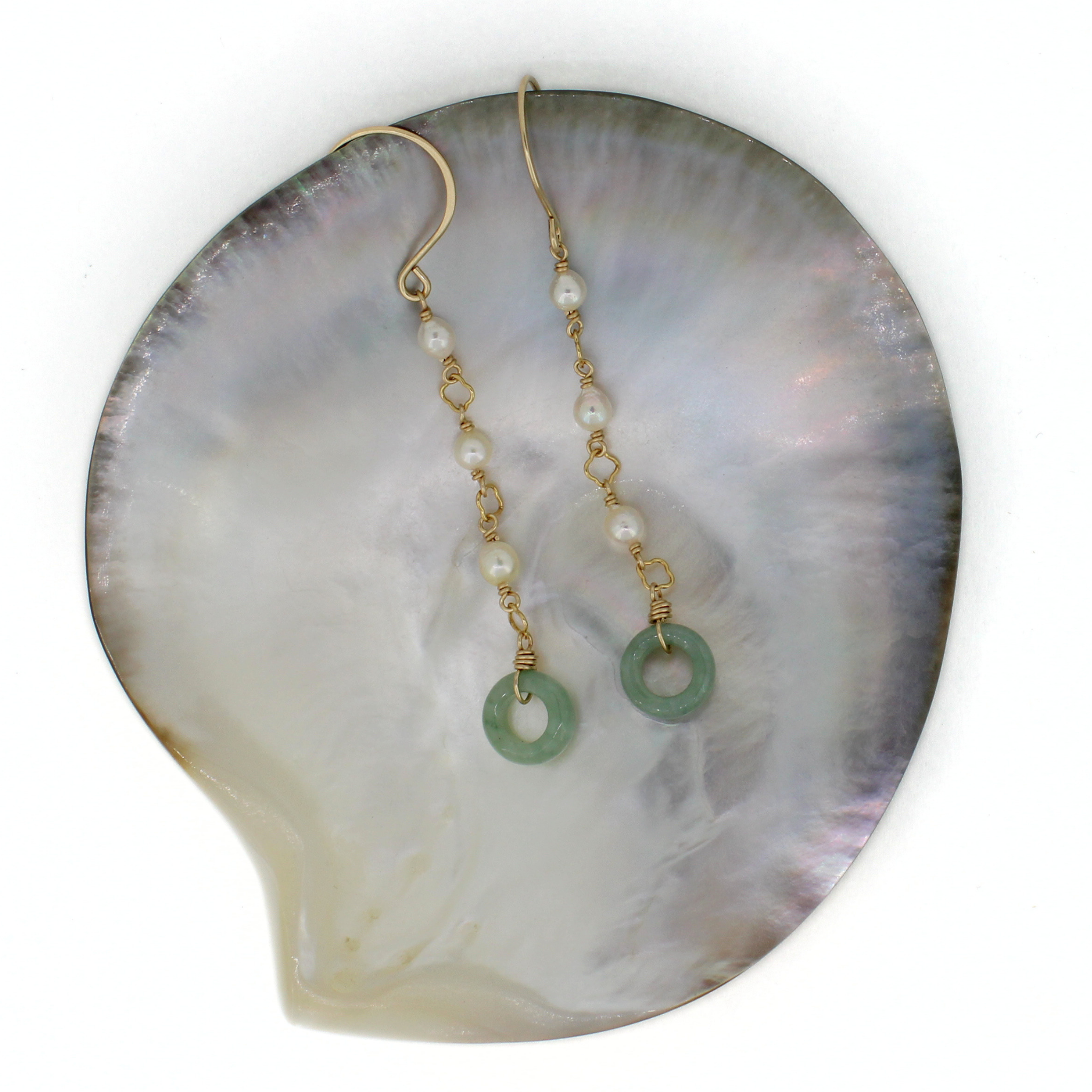 A pair of gold dangle earrings with small white pearls and circular green jade charms, displayed on a large iridescent shell against a white background.