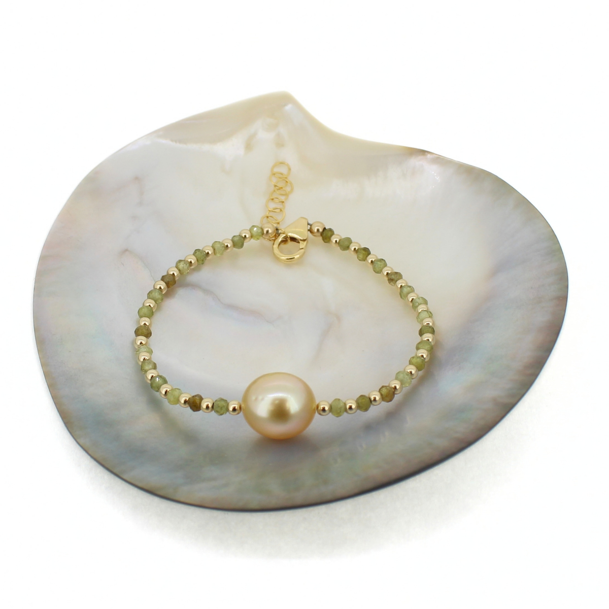 A delicate gold beaded bracelet with a large round pearl and small faceted green beads, displayed on a white clam shell.