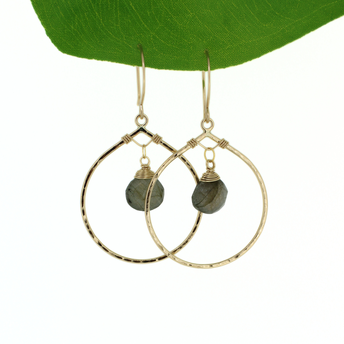 A pair of gold hoop drop earrings with small faceted dark gray labradorite stones, hanging against a white background with a green leaf at the top.